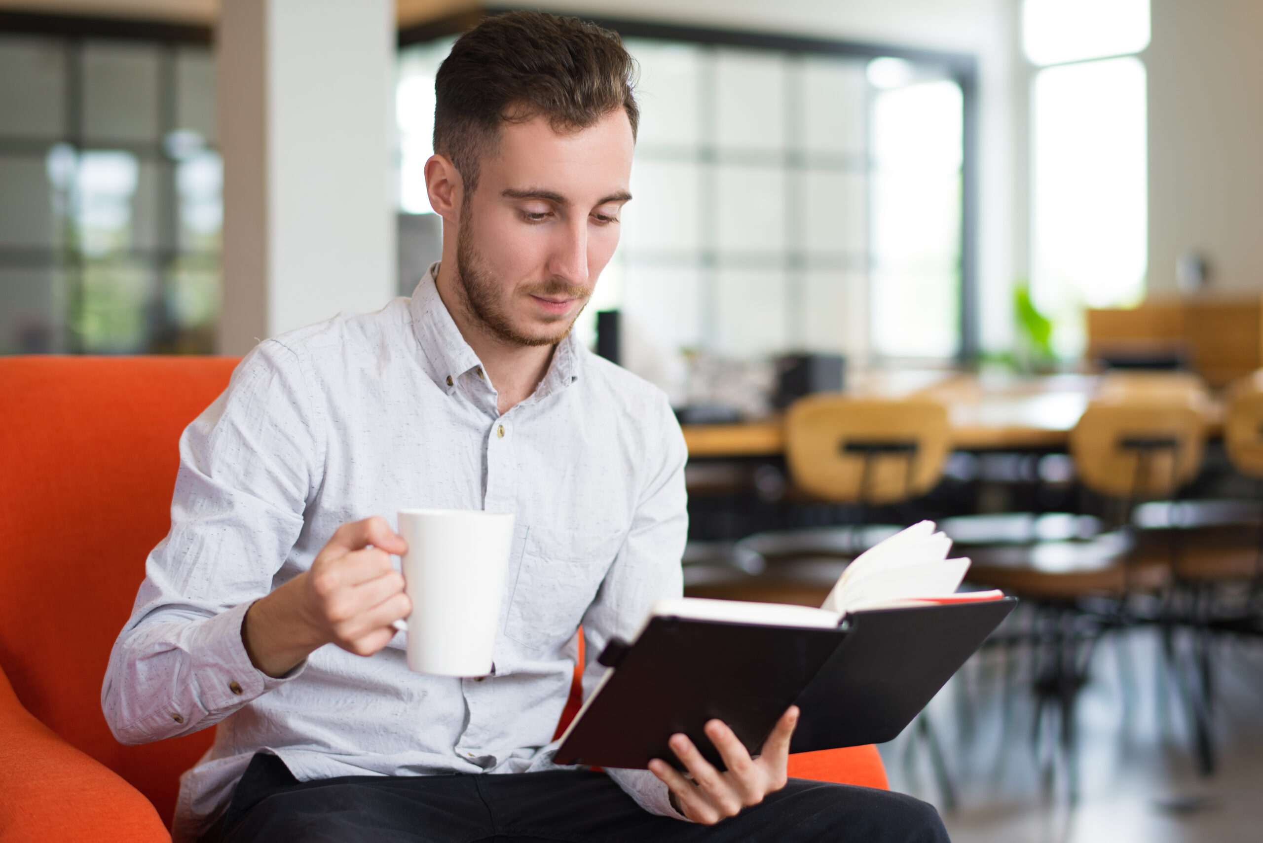 Homem lendo livro, sentado no sofá e tomando café